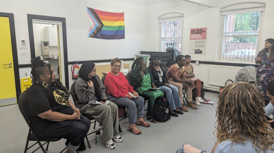 Image of The image shows seven seated people facing an audience. The wall behind them has a rainbow colour pride progress flag on it.