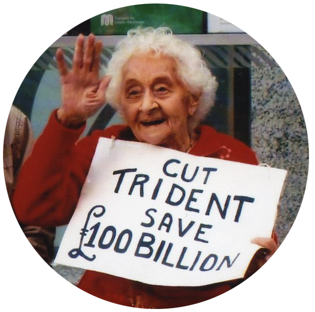 Colour photograph of a white female, Betty Tebbs, holding a sign with text: 'Cut Trident Save £100 Billion'.