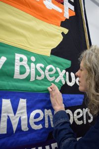 A white woman (PHM Conservation Manager Jenny Van Enckevort) looking up close at a rainbow coloured banner including partially visible white text: 'Bisexual' and 'Members'.