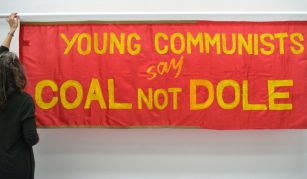 Image of A landscape rectangular red banner with yellow text: ‘Young Communists say Coal Not Dole’ with a woman (PHM Conservation Manager Jenny Van Enckevort) standing on the left, reaching up to adjust the top of the banner on its hanging pole.