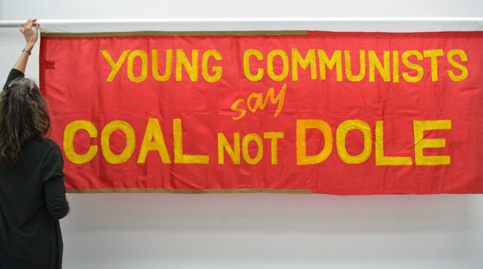 Image of A landscape rectangular red banner with yellow text: ‘Young Communists say Coal Not Dole’ with a woman (PHM Conservation Manager Jenny Van Enckevort) standing on the left, reaching up to adjust the top of the banner on its hanging pole.