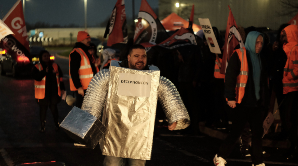 Image of Photograph showing a person in a silver robot costume with 'Deceptioncon' written on the front, and people in the background wearing orange high-vis jackets and waving GMB union flags.