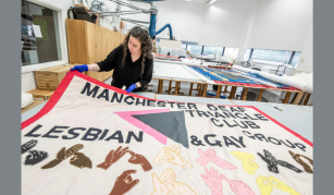 Image of Conservator with blue gloves holding up a textile banner featuring text: 'Manchester Deaf Triangle Club, Lesbian & Gay Group', and different coloured hands doing sign language.