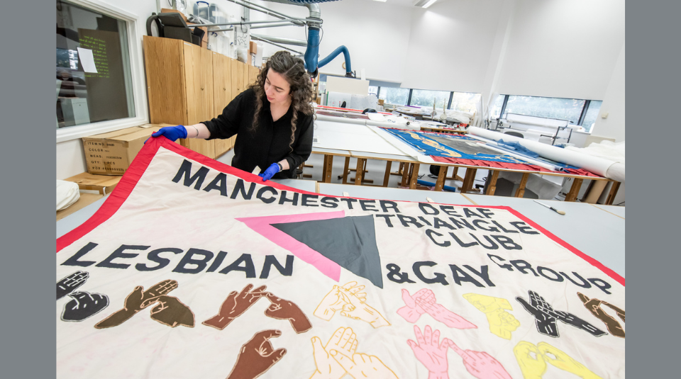 Image of Conservator with blue gloves holding up a textile banner featuring text: 'Manchester Deaf Triangle Club, Lesbian & Gay Group', and different coloured hands doing sign language.