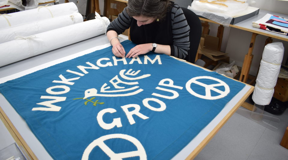 Image of A white woman (PHM Senior Conservator Kloe Rumsey) working on a large blue banner laid out on a table with white text: 'Wokingham Peace Group'.