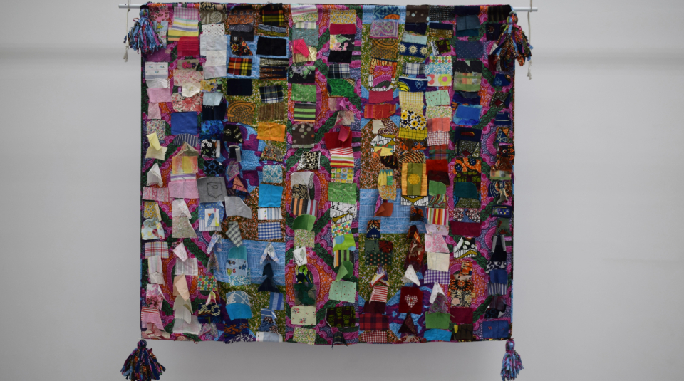 Image of A square shaped quilt made up of hundreds of small scraps of material. Each scrap has a different pattern or colour. The four corners of the quilt have tassels, that have been made out of similar offcuts of material.