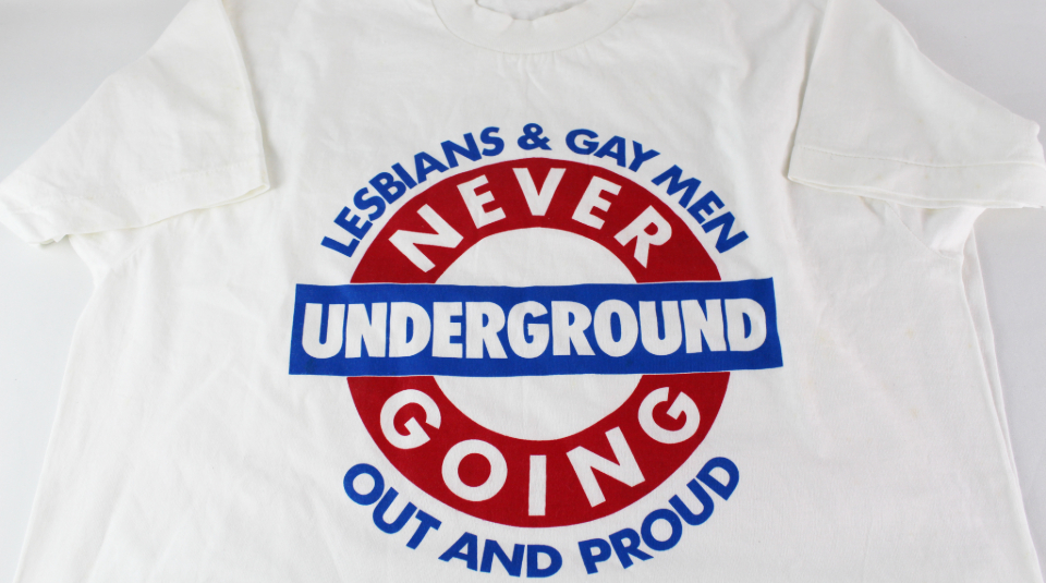 Image of white t-shirt with blue and red London Underground style logo with text: 'Lesbians & Gay Men, Never Going Underground, Out And Proud'.