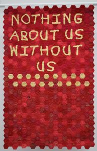 A red textile banner made up of stitched hexagons, with gold text: 'Nothing About Us Without Us'.