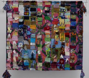 A square shaped quilt made up of hundreds of small scraps of material. Each scrap has a different pattern or colour. The four corners of quilt have tassels, that have been made out of similar offcuts of material. 