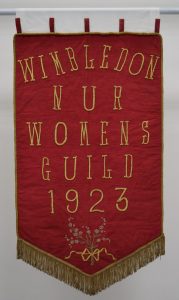 A shield shaped red banner with gold trim around the edges The central design is hand embroidered gold lettering that reads ‘WIMBLEDON NUR WOMENS GUILD 1923’.