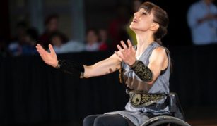 Image of A person in a wheelchair with decorative armbands and a belt is performing, with arms raised and looking upwards, likely in a public or stage setting with an audience in the background.
