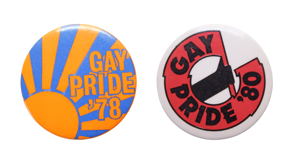 Round blue badge reading ’Gay Pride '78’ with an orange sun with rays and a white round badge with ‘GAY PRIDE '80’ in a red flag wrapping around the edge of the badge.