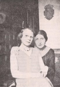 A black and white photograph of Charlotte Charlaque and Toni Ebel. Toni in a pale dress sits on Charlotte’s lap – both are looking at the camera.