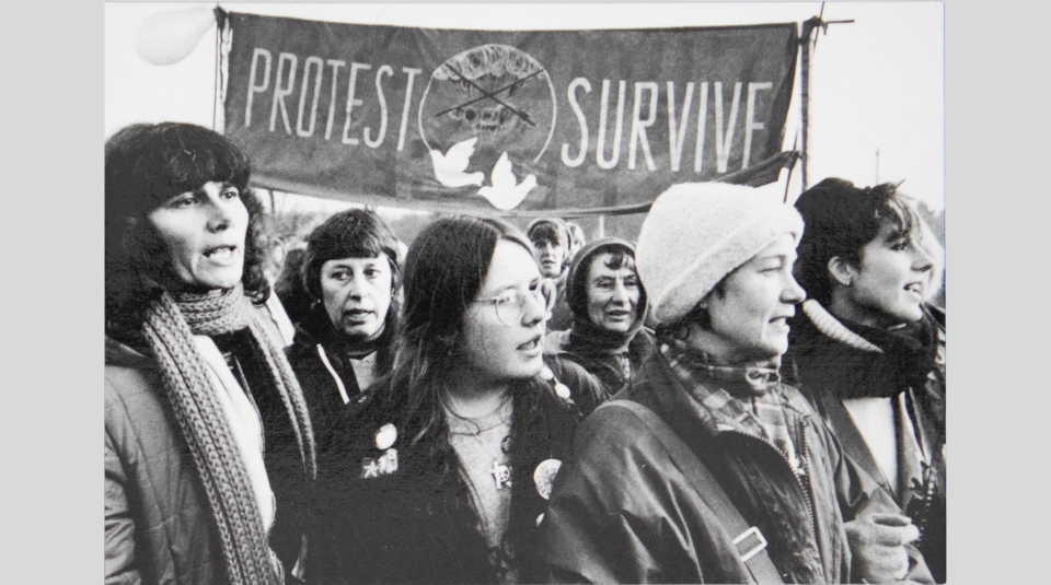Image of Black and white photograph postcard, a group of people outside with “Protest - Survive” banner in the background, featuring two white doves in the middle.