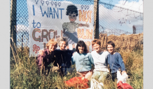 Image of Colour photograph of five women with their arms around each other, sat in long grass in front of a fence with barbed wire at the top. Fabric banner on the fence features a toddler and text 