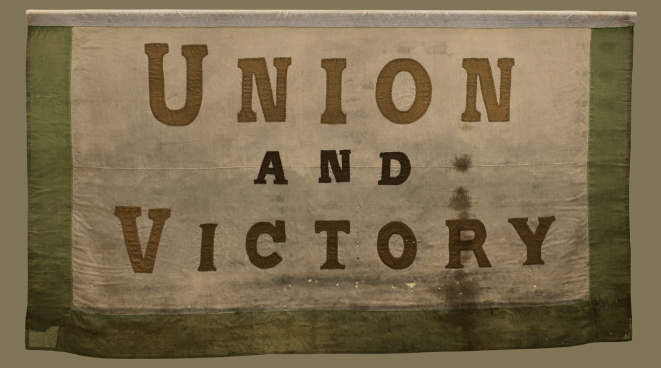 Image of A beige rectangular banner with a dark olive green border. The words ‘UNION AND VICTORY’ are prominently displayed in large golden brown letters