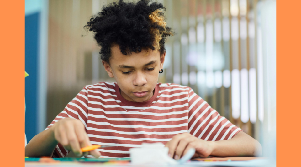 Image of A child seated at a table holding scissors and paper
