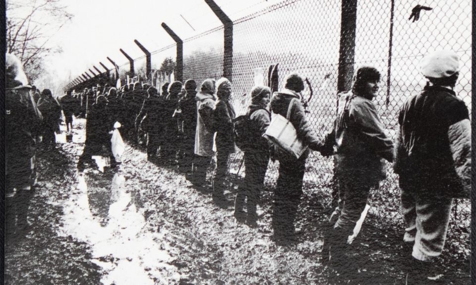 Black and white photograph of women holding hands in a long line. They are standing along the chain-link fence topped with barbed wire at RAF base Greenham Common. 