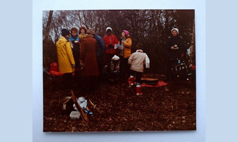 Colour photograph of a group of women and a child talking in small groups in the woods at Greenham Common Peace Camp. They wear raincoats and the trees are bare.
