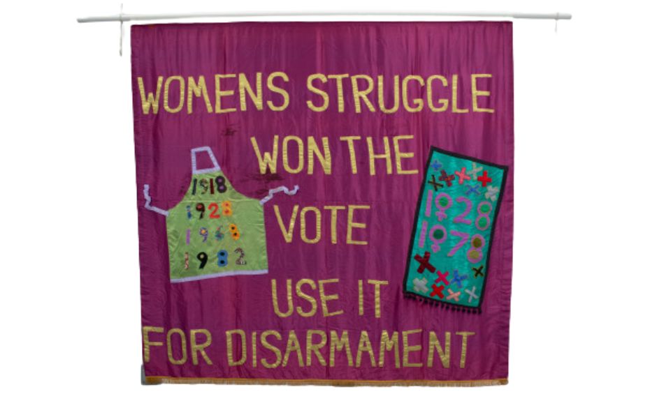 a purple banner with the words ‘WOMENS STRUGGLE WON THE VOTE USE IT FOR DISARMAMENT’ and two images. One an apron, with the years ‘1918, 1928, 1968, 1982’. The second a banner with the years ‘1928, 1978.’