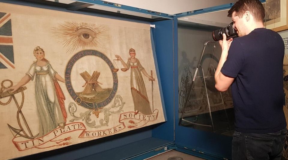 Image of A person looking into a camera on a tripod, photographing a banner on display at People's History Museum. The banner is the Tin Plate Workers Society banner from 1821.