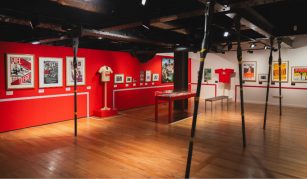 Image of A photograph of an exhibition space with red painted walls, with displays including framed posters, t-shirts, and photographs.