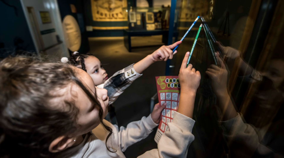 Image of 2 children pointing at a display case with a pencil and a banner bingo card in their hands