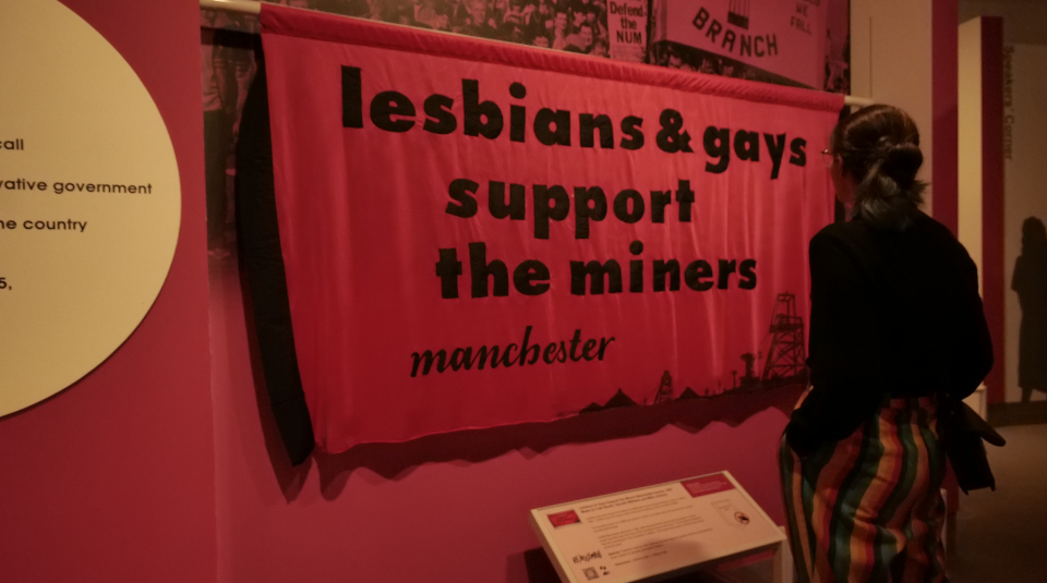 Image of A person looking at a pink banner which reads 'lesbians & gays support the miners - Manchester'