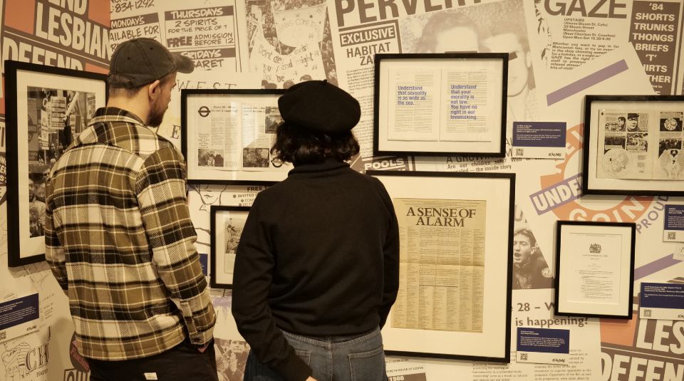 Two people stood looking at framed archive materials on a collage backdrop including newspapers and leaflets.
