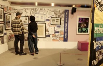 Image of Two people stood looking at framed archive materials on a collage backdrop including newspapers and leaflets.