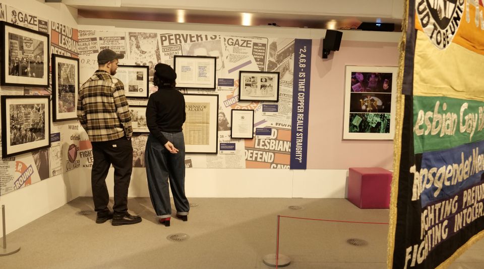 Image of Two people stood looking at framed archive materials on a collage backdrop including newspapers and leaflets.