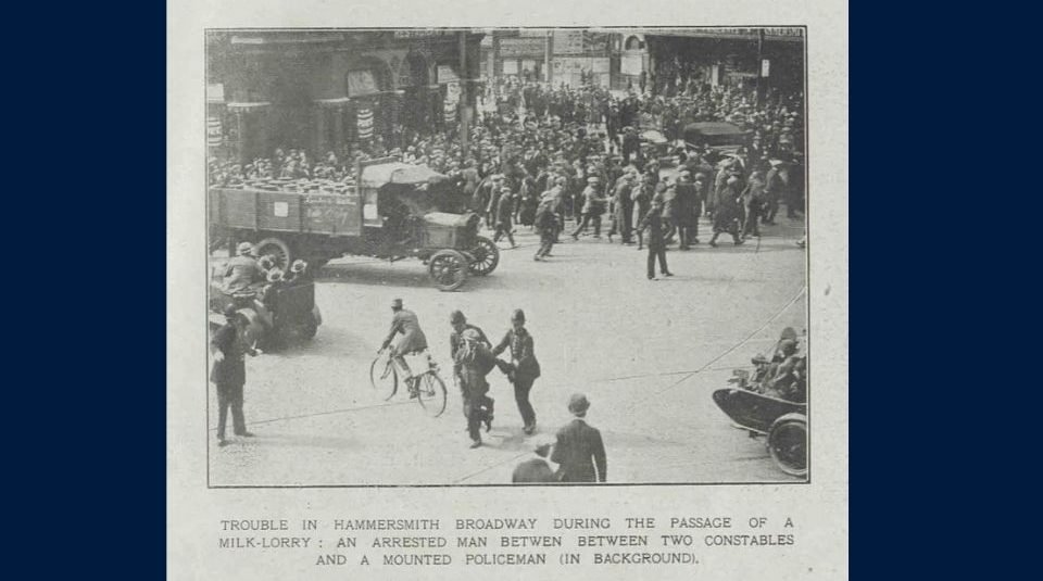 Image of Historic black-and-white photograph of a crowded street in Hammersmith Broadway, with police arresting a man during the passage of a milk lorry, surrounded by onlookers, vehicles, and a mounted policeman in the background.