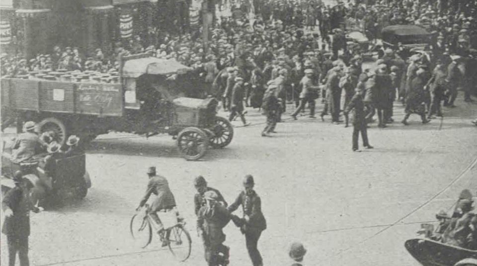 Image of Historic black-and-white photograph of a crowded street in Hammersmith Broadway, with police arresting a man during the passage of a milk lorry, surrounded by onlookers, vehicles, and a mounted policeman in the background.