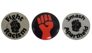 Image of Left to right: a white badge with a black raised fist with text reading: 'Fight Racism', a black badge with a red raised fist, and a white badge with a black raised fist with text reading: 'Smash Apartheid'.