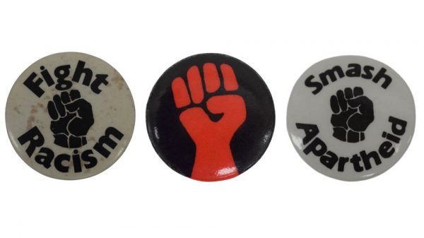 Image of Left to right: a white badge with a black raised fist with text reading: 'Fight Racism', a black badge with a red raised fist, and a white badge with a black raised fist with text reading: 'Smash Apartheid'.