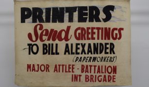 Image of Photograph of a cream banner with bold black and red text reading 'PRINTERS Send GREETINGS TO BILL ALEXANDER (PAPERWORKERS) MAJOR ATTLEE - BATTALION INT. BRIGADE.'