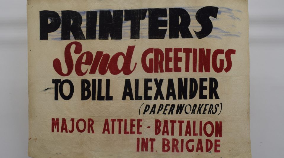 Image of Photograph of a cream banner with bold black and red text reading 'PRINTERS Send GREETINGS TO BILL ALEXANDER (PAPERWORKERS) MAJOR ATTLEE - BATTALION INT. BRIGADE.'
