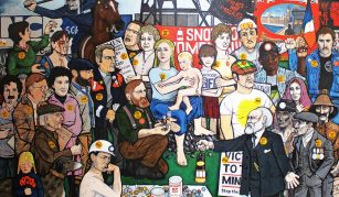 Image of A colorful mural depicting a diverse group of people, including miners children, and police, gathered together. Various signs, badges with text which includes Coal Not Dole and Mines Not Missiles