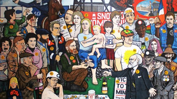 Image of A colourful mural depicting a diverse group of people, including miners, children, and police gathered together, along with various signs and badges with text including Coal Not Dole and Mines Not Missiles.