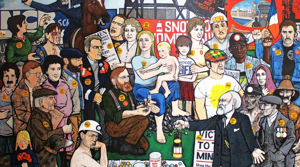 Image of A colourful mural depicting a diverse group of people, including miners, children, and police gathered together, along with various signs and badges with text including Coal Not Dole and Mines Not Missiles.