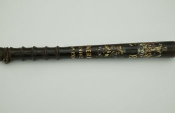 Image of Photograph of a vintage black wooden truncheon with a textured grip and faded gold lettering along its length. The text includes May 1926.