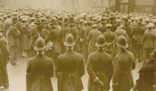 Image of Photograph of a dense crowd of men surrounded by police, amongst a backdrop of buildings. The crowd is facing away from the camera.