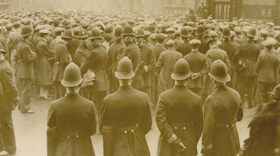 Image of Photograph of a dense crowd of men surrounded by police, amongst a backdrop of buildings. The crowd is facing away from the camera.