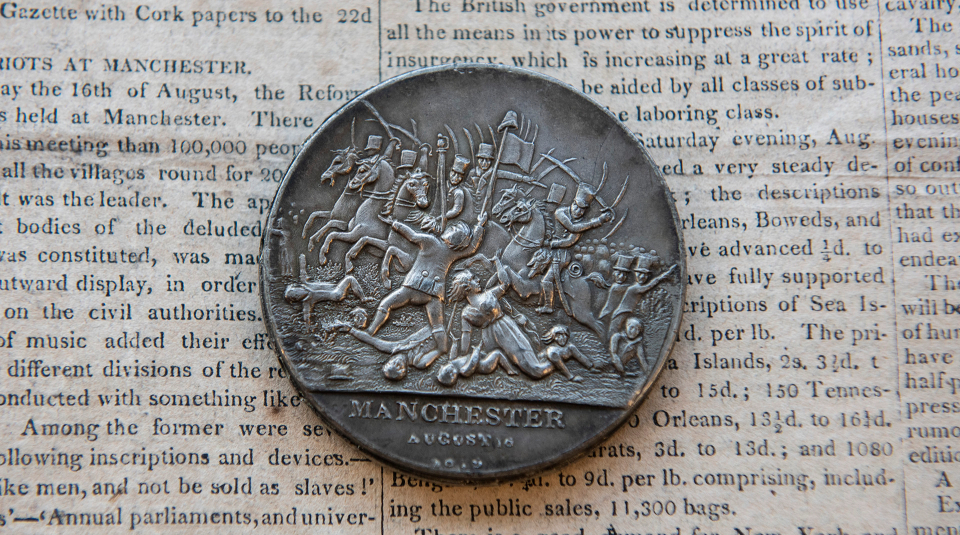 Image of A silver medal featuring an image of men in uniform on horseback striking swords on people below, and text 'August 16', placed on a newspaper page of text.