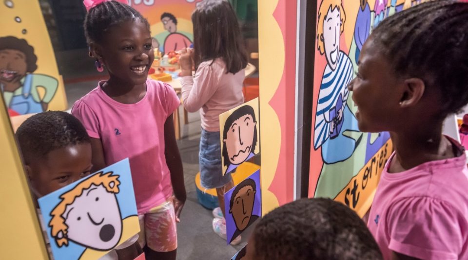 Image of Photograph showing children interacting with a colorful mirror installation featuring cartoon faces expressing different emotions
