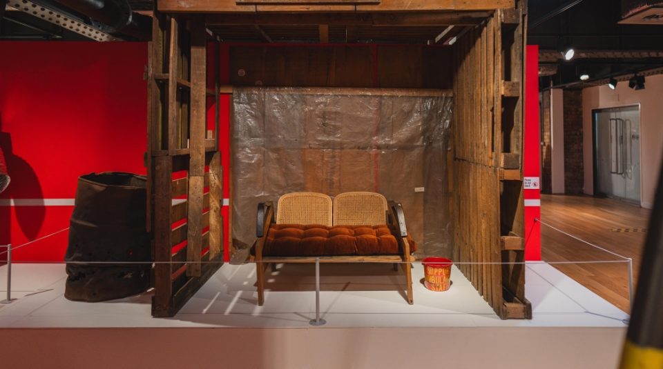 Image of A wooden shelter.  Inside is a two-seater bench with cushions and a bucket labelled   ‘SUPPORT AMBULANCE STAFF’ Next to shelter is a rusted metal drum.