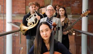 Image of Photograph of a group of five musicians standing indoors against a brick wall, each holding different instruments including a cello, French horn, and bass guitar.