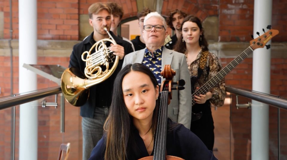 Image of Photograph of a group of five musicians standing indoors against a brick wall, each holding different instruments including a cello, French horn, and bass guitar.