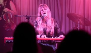 Image of Photograph of a live music performance featuring a musician playing a keyboard and singing into a microphone, with a violinist partially visible on the left and a drum set on the right. The scene is illuminated with pink stage lighting, and silhouettes of audience members are visible in the foreground.