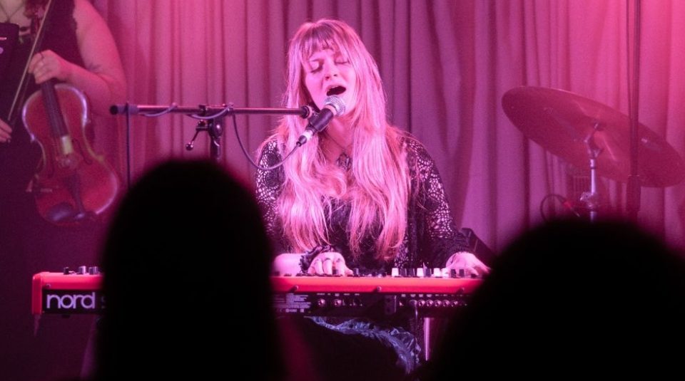 Image of Photograph of a live music performance featuring a musician playing a keyboard and singing into a microphone, with a violinist partially visible on the left and a drum set on the right. The scene is illuminated with pink stage lighting, and silhouettes of audience members are visible in the foreground.